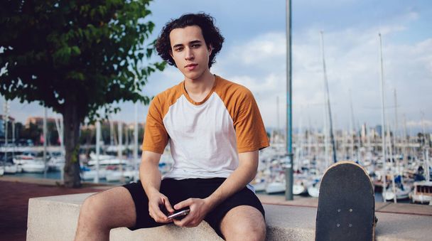 Young curly haired male hipster in trendy casual wear sitting on rocky fence with mobile phone skateboard and looking at camera in Barcelona - Foto, afbeelding
