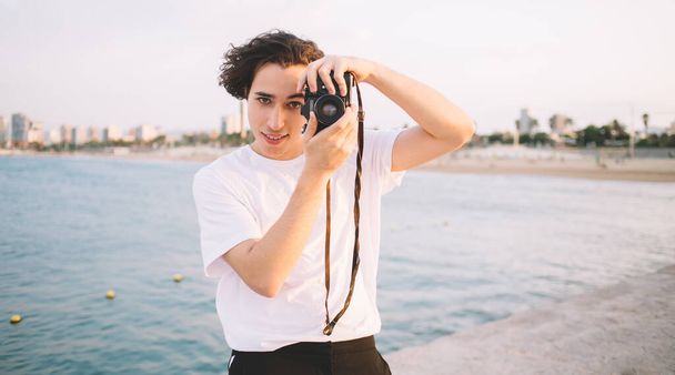 Joyful young hipster guy using photo camera standing in front of clear sea water and looking at camera while enjoying sunny day - Fotoğraf, Görsel