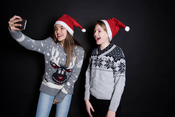 Vrolijk Kerstfeest en Gelukkig Nieuwjaar! mooie broer en zus thuis nemen een selfie met mobiele telefoon. gelukkig kinderen dragen santa hoed op zwart achtergrond. - Foto, afbeelding