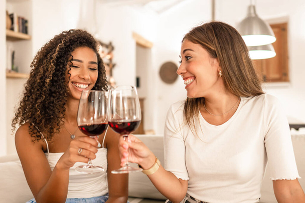 Две молодые женщины смешанной расы празднуют дома тост с красными бокалами вина - улыбаясь латиноамериканская кудрявая брюнетка звон с ее лучшим другом сидя на диване в ее современной элегантной гостиной - Фото, изображение
