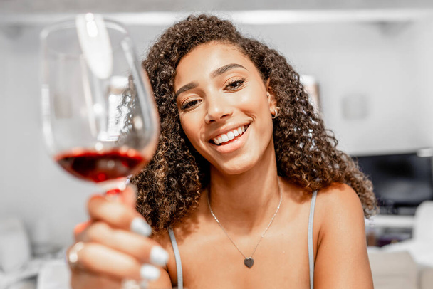 幸せな若いヒスパニック系の巻きブルネットは、ビデオ通話会議でカメラを見て赤ワイングラスで乾杯-美しい黒の女性は、新しいソーシャルネットワークとオンラインチャットリビングルームに座っています - 写真・画像