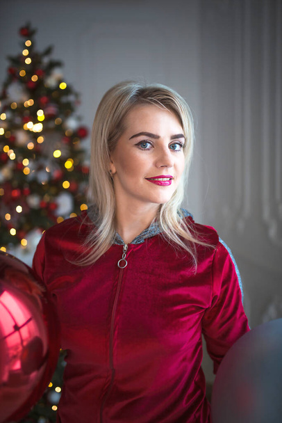 Young blonde woman in a red tracksuit on Christmas decorations, on the background of a Christmas tree. Big balls, lights, garlands, and home decor. Sports lifestyle. New Year. Make-up for the holiday - Photo, Image