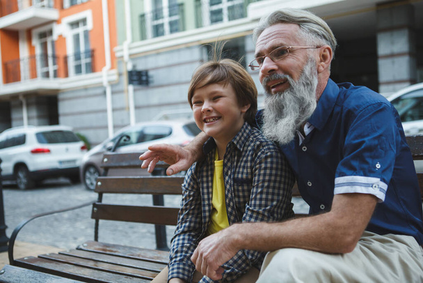 Happy senior man hugging his grandson, sitting together on the bench at city center - Fotoğraf, Görsel