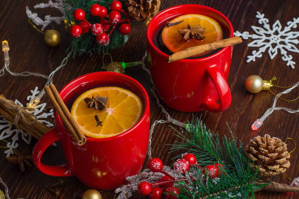 Ароматическое, горячее и вкусное глинтвейн с апельсином, корицей, звездным анисом и гвоздикой в красных чашках с рождественским декором. Ощущение праздника и тепла в доме. - Фото, изображение