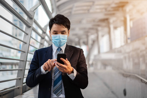 business man using a smartphone and wearing medical mask during coronavirus (covid-19) pandemic in the city - 写真・画像