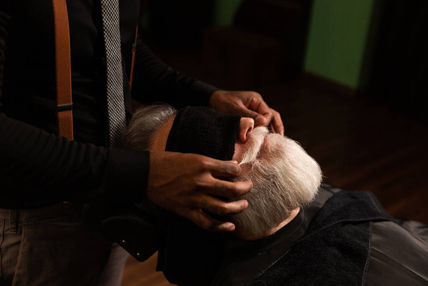 Las manos del barbero afroamericano peinan, ajustan y enceran el bigote de un hombre mayor con una barba gris, el cliente usa una toalla negra sobre sus ojos - Foto, Imagen