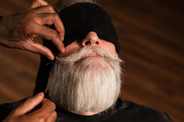 African-American barber's hands comb, adjust and wax the mustache of an older man with a gray beard, the customer wears a black towel over his eyes - Photo, Image