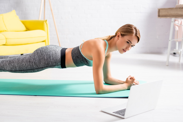 Blonde sportswoman standing in plank near laptop on blurred foreground on floor  - Foto, immagini