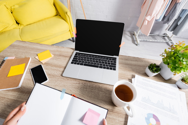 Laptop és okostelefon üres képernyőn közel csésze tea, papírok és szabadúszó gazdaság notebook elmosódott előtérben otthon - Fotó, kép