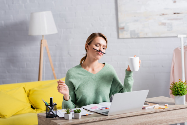 Freelancer holding cup and pen near lips while working with papers and laptop at home  - Фото, изображение