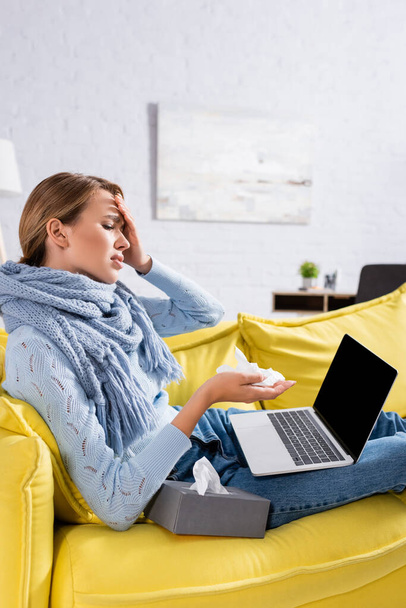 Femme malade avec écharpe tenant la serviette et regardant l'ordinateur portable avec écran blanc sur le canapé  - Photo, image