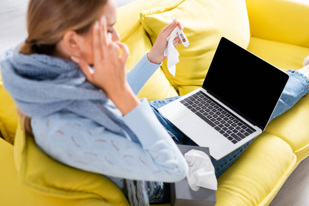 Portátil con pantalla en blanco cerca de la mujer enferma con termómetro y servilletas en primer plano borroso en el sofá  - Foto, Imagen