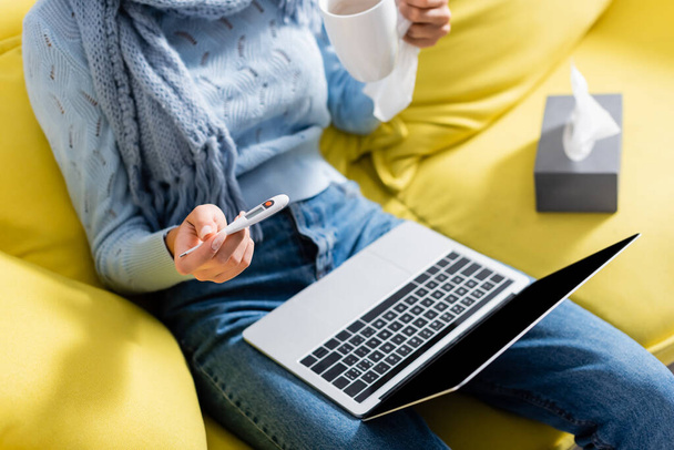 Cropped view of freelancer holding thermometer and cup near laptop and napkins on blurred background  - Photo, image