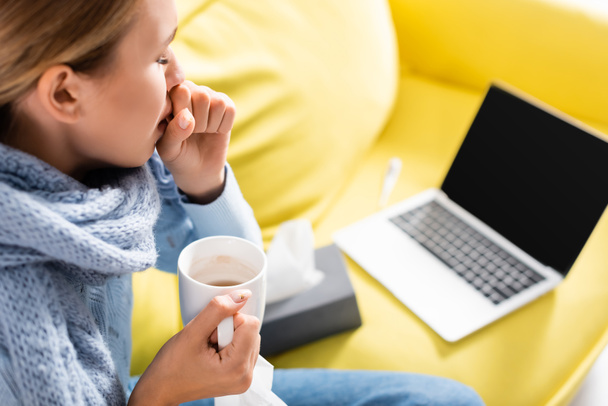 Άρρωστη γυναίκα βήχει κρατώντας κύπελλο κοντά στο laptop και χαρτοπετσέτες σε θολή φόντο  - Φωτογραφία, εικόνα