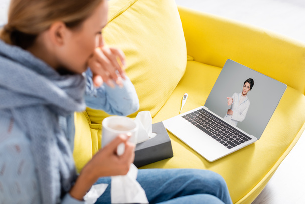 Doctor pointing with hand on screen of laptop near sick woman holding cup during video call on blurred foreground  - Foto, imagen