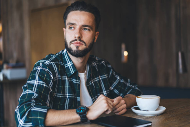Thoughtful bearded young male in checkered shirt looking away while drinking coffee at daytime in modern cafe with wooden furniture - Foto, Bild