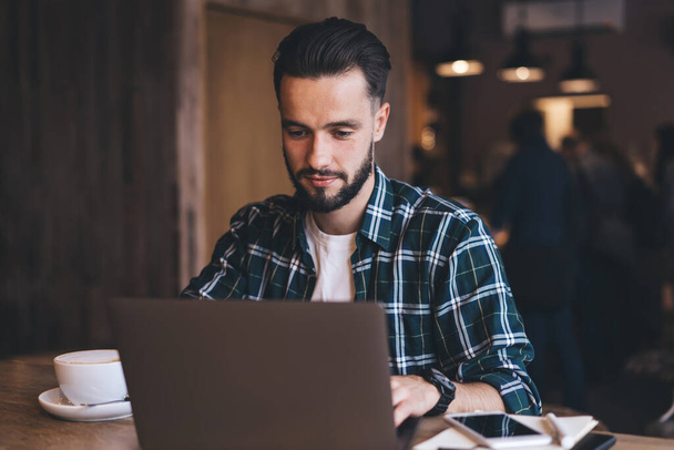 Concentrated bearded male freelancer in checkered shirt using laptop while sitting at table with cup of coffee and smartphone in loft cafeteria - 写真・画像