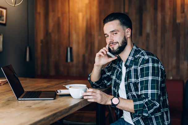 Bearded male blogger in casual clothing and wristwatch answering on phone call while enjoying cup of coffee in creative caferetia - Fotografie, Obrázek