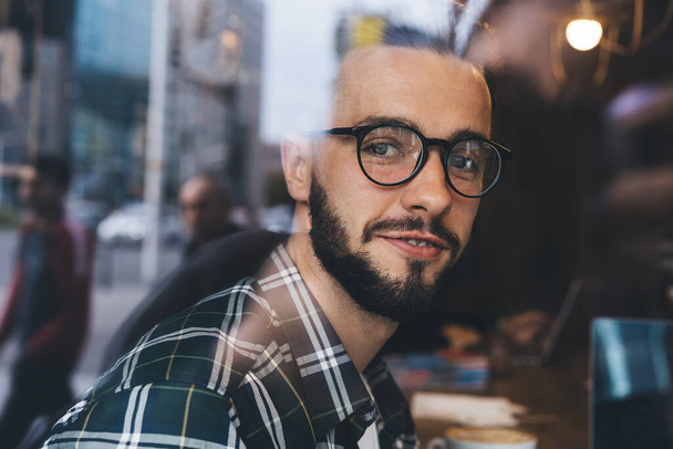 Through glass view of bearded young guy in green checkered shirt and glasses sitting in cafe while smiling and looking at camera - Zdjęcie, obraz