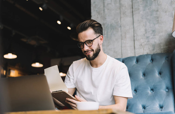 Low angle of happy bearded male in casual outfit and eyeglasses flipping page of clipboard and reading article on laptop while smiling and sitting on couch in cafe - Zdjęcie, obraz