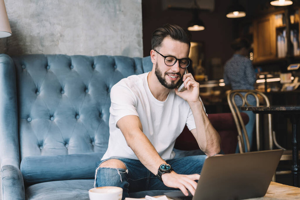 Satisfied young male with beard wearing informal outfit and glasses browsing laptop and talking on mobile phone while sitting at table in cafe - Foto, imagen