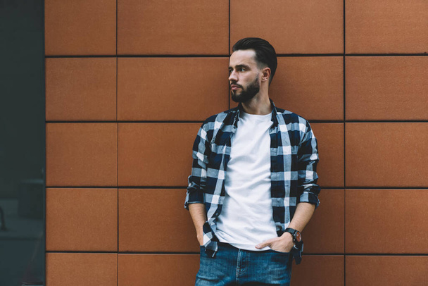 Pensive young bearded stylish guy leaning on city building wall with hands in pockets of jeans looking away standing outside alone - Photo, Image