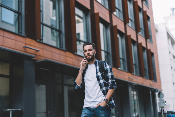 Low angle of modern hipster male in casual wear with backpack walking down city street while talking on smartphone and looking at  camera - 写真・画像