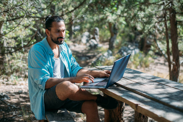 Männliche Touristen arbeiten draußen auf einem Campingplatz an einem Laptop. Junger Mann, Freiberufler, der bei einer Bergwanderung im Freien am Laptop arbeitet. Die Fähigkeit, von jedem Ort der Welt aus zu arbeiten. - Foto, Bild