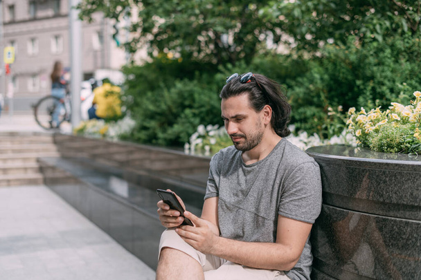 A man sits on a bench with a phone on the street of a big city. Young white handsome guy walks on foot in the metropolis, rests and communicates on a smartphone on a green city street - Фото, изображение