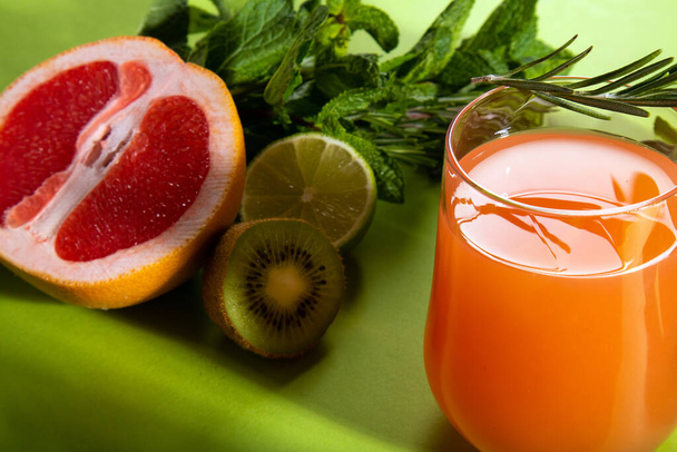 Composizione di bicchieri con succo d'arancia, taglio rosso arancio, lime, kiwi, rosmarino e foglie di menta su fondo verde chiaro - Foto, immagini