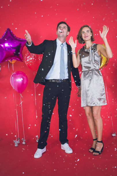 Mladý pár je šťastný mezi konfety, které padají vepředu. Vedle nich jsou barevné balónky a červené pozadí v konceptu Nového roku a Vánoc. - Fotografie, Obrázek