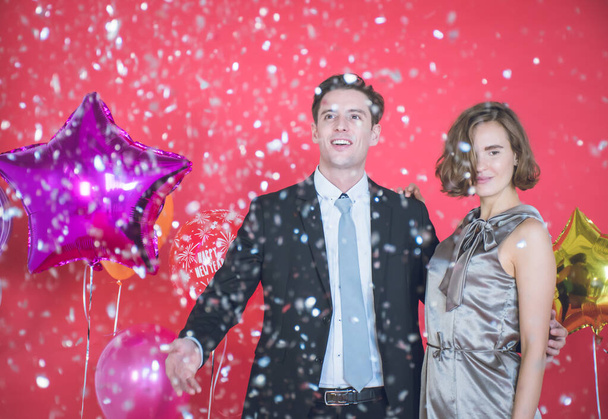 O jovem casal está feliz entre os confetes que caem na frente. Há balões coloridos ao lado deles e o fundo vermelho no conceito de Ano Novo e dia de Natal. - Foto, Imagem