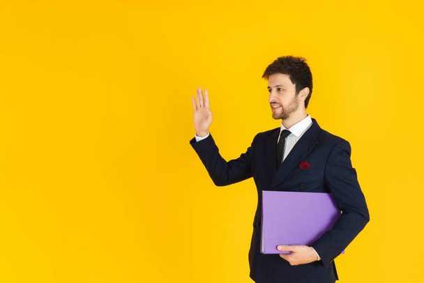 Ein junger Geschäftsmann im Anzug hält eine Akte mit Dokumenten in der Hand und winkt auf dem gelben Hintergrund. Geschäftsimage für Banner. - Foto, Bild