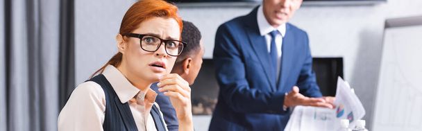 Scared redhead businesswoman looking at camera with blurred multicultural colleagues on background, banner - Photo, Image