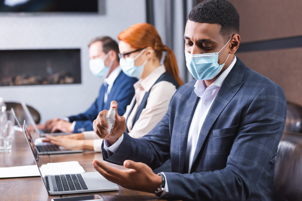 Tıbbi maskeli Afrikalı Amerikalı işadamı iş yerindeki bulanık arka planda dezenfektan kullanıyor. - Fotoğraf, Görsel