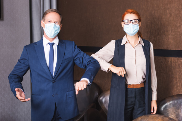 Orvosi maszkos üzletemberek nézik a kamerát, miközben könyökkel üdvözlik egymást a tárgyalóban. - Fotó, kép