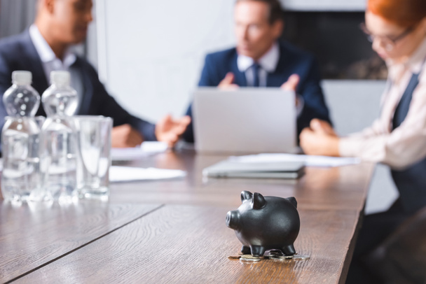 Small black piggy bank with coins on table with blurred multicultural businesspeople on background - Photo, Image