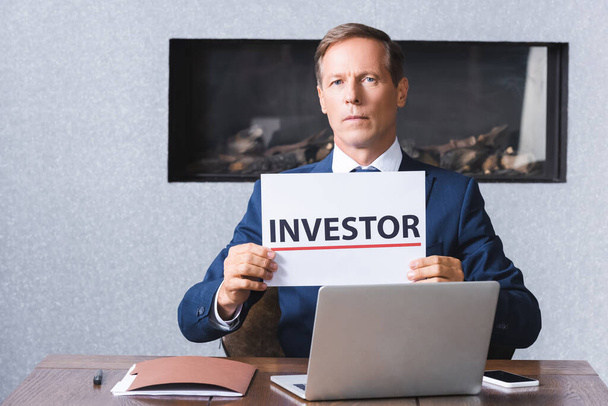 Серьезный бизнесмен смотрит в камеру и держит табличку с надписью инвестора на рабочем месте с цифровыми устройствами в конференц-зале - Фото, изображение