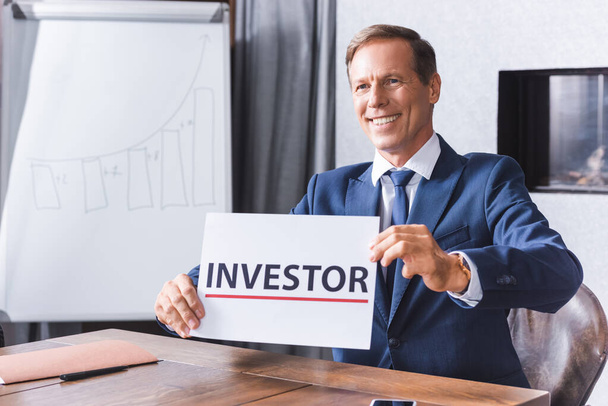 Smiling businessman looking away while showing sign with investor lettering at workplace in meeting room - Valokuva, kuva