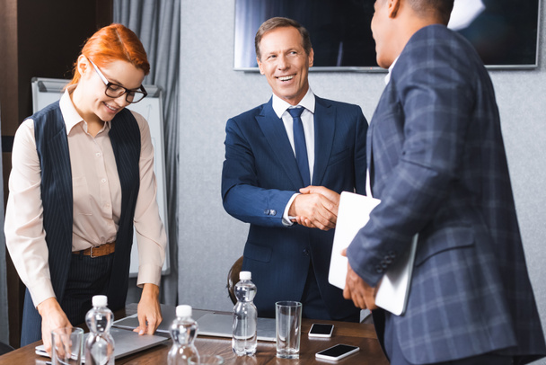 Lächelnder Geschäftsmann beim Händeschütteln mit afrikanisch-amerikanischem Kollegen, während er neben Geschäftsfrau im Besprechungsraum steht - Foto, Bild