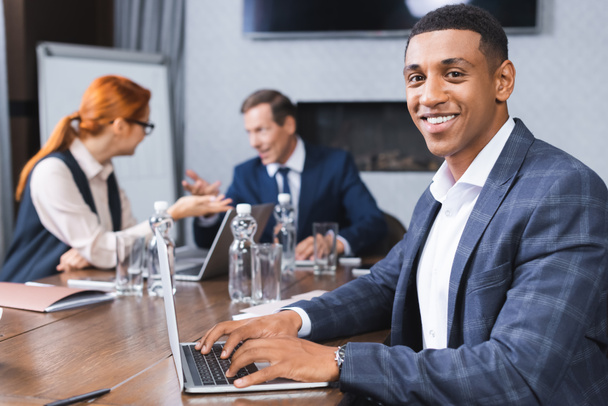 Happy african american businessman looking at camera while typing on laptop with blurred colleagues talking on background - Foto, imagen