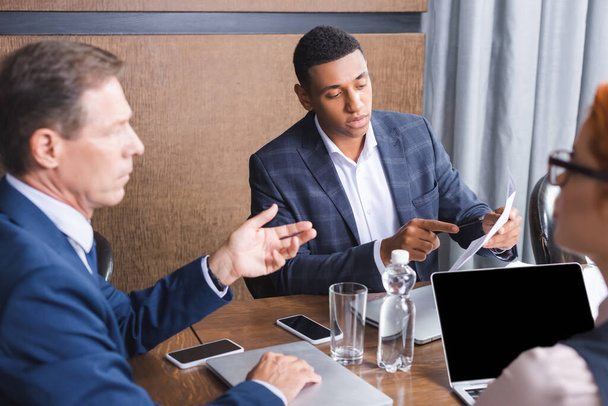 Africký americký podnikatel ukazuje prstem na papír, zatímco sedí na pracovišti v blízkosti rozmazaných kolegů na popředí - Fotografie, Obrázek