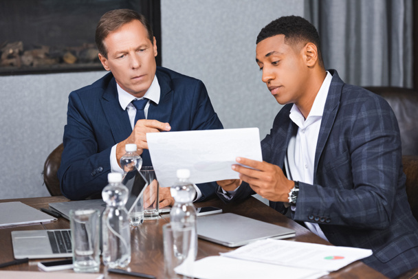 Des hommes d'affaires multiculturels regardant du papier assis sur le lieu de travail avec des appareils numériques dans la salle de réunion - Photo, image