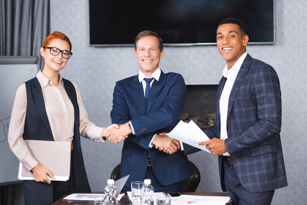 Happy businessman looking at camera while shaking hands with multicultural colleagues near workplace in boardroom - Photo, Image