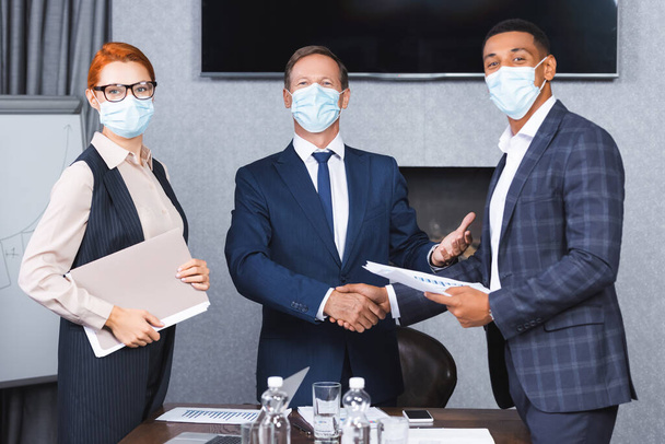 Multikulturelle Geschäftsleute in medizinischen Masken schütteln einander die Hände, während sie neben Geschäftsfrau im Sitzungssaal stehen - Foto, Bild