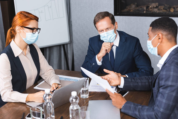 Empresarios multiculturales en máscaras médicas hablando mientras están sentados en el lugar de trabajo con dispositivos y documentos digitales - Foto, Imagen