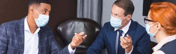 Multicultural businesspeople in medical masks gesturing during discussion near female colleague in boardroom, banner - Photo, Image