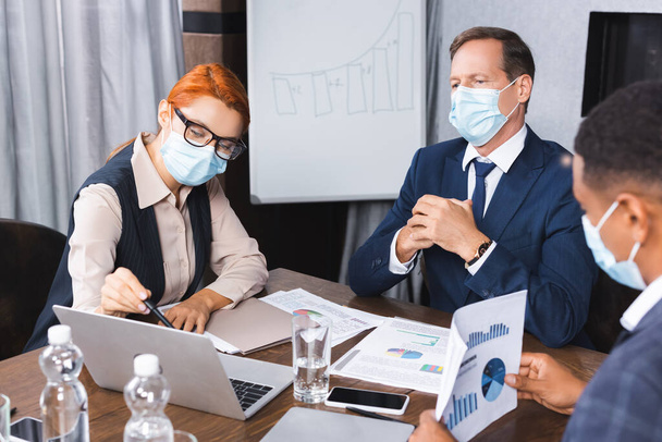 Geschäftsfrau in medizinischer Maske zeigt mit Stift auf Laptop in der Nähe von Investor mit verschwommenem afrikanisch-amerikanischem Kollegen im Vordergrund - Foto, Bild