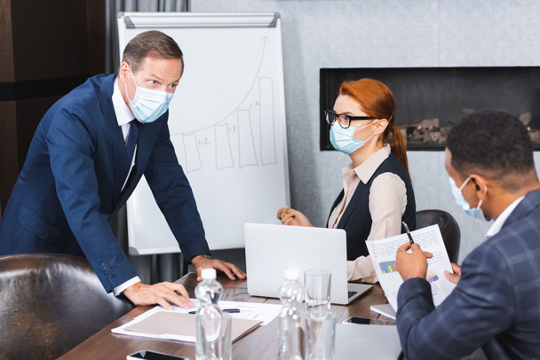 Businessman in medical mask looking at african american colleague while leaning on workplace in meeting room - Photo, Image