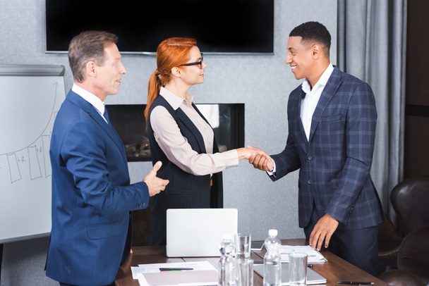 Χαμογελώντας πολυπολιτισμικοί επιχειρηματίες σφίγγουν το χέρι ο ένας στον άλλο κοντά στο συνάδελφο στο χώρο εργασίας - Φωτογραφία, εικόνα
